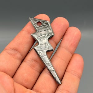 Könnte beinhalten: Ein Metall-Blitz-Anhänger mit einem Loch für eine Kette. Der Anhänger hat ein einzigartiges Muster aus hellen und dunklen Wirbeln, das ihm ein strukturiertes Aussehen verleiht. Das Design ist eine stilisierte Darstellung eines Blitzes.