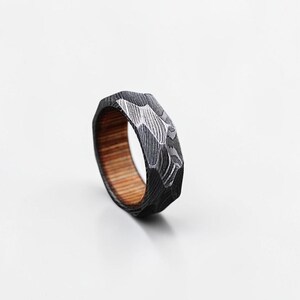 Pode incluir: Um anel facetado com um exterior escuro e texturizado e um interior de madeira castanha clara. O exterior do anel tem um design padronizado com detalhes prateados. O anel está sobre um fundo branco.