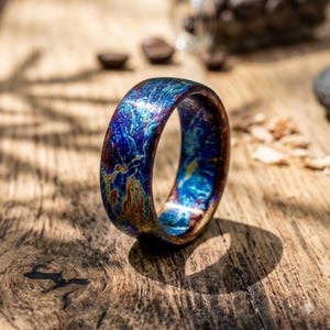 May include: A ring featuring a polished surface with a swirling, iridescent pattern of deep blue, purple, and metallic gold tones. The band has a smooth, rounded exterior and a glossy finish that reflects light across its colorful, marbled texture.