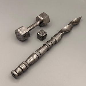 May include: A set of three metal objects: a small dumbbell, a die, and a pen. The dumbbell has a bar with two hexagonal weights. The die is a small cube with dots. The pen has a twisted design.
