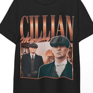 Peut inclure: T-shirt noir avec une image de style vintage de Cillian Murphy. Le design comprend un portrait de l'acteur et le nom "Cillian Murphy" en cuivre. Le t-shirt est suspendu à un cintre en bois.