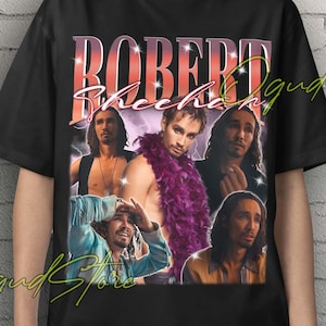 May include: Black Unisex T-shirt featuring a collage of images and the name "ROBERT Sheehan" in a retro style. The design includes multiple portraits of the actor, with a purple feather boa. The shirt has a crew neck.