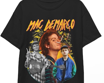Op de afbeelding: Zwart t-shirt met een afbeelding van Mac DeMarco en de tekst "MAC DEMARGO" in geel en oranje. De afbeelding bevat drie afbeeldingen van de muzikant. Het t-shirt hangt aan een houten hanger.