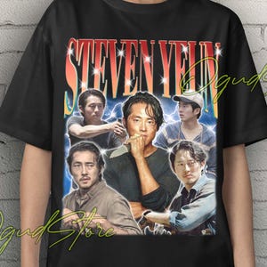 Puede incluir: Camiseta unisex negra con un diseño gráfico retro de Steven Yeun. El diseño incluye múltiples imágenes del actor y el texto "STEVEN YEUN" en una fuente retro. La camiseta la lleva una persona.