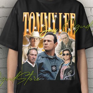 May include: Black Unisex T-shirt featuring a vintage-style graphic of Tommy Lee Jones with the text "Tommy Lee Jones" in a gold font. The shirt has a crew neck and short sleeves. The shirt is worn by a person.