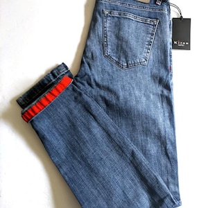 May include: Blue denim jeans with a red stripe detail on the folded cuff. The jeans feature a classic five-pocket design, a leather patch, and a black tag with the brand name "Kiton".