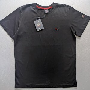 May include: A black short-sleeved t-shirt featuring a small embroidered shark logo on the left chest and a matching patch on the left sleeve. The shirt includes a red inner neck trim and a black hang tag with the Paul & Shark logo attached.