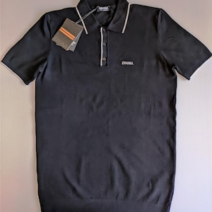 Könnte beinhalten: Ein schwarzes kurzärmeliges Poloshirt mit einem Kragen mit weißem Rand, einer Knopfleiste mit drei Knöpfen und weißer Paspelierung sowie dem in Weiß auf die Brust gestickten Wort ZEGNA. Ein Markenetikett ist mit einem schwarzen Band am Kragen befestigt.