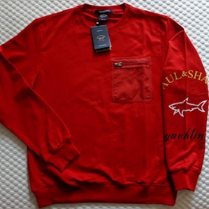 May include: A bright red long-sleeve sweatshirt featuring a crew neckline, a zippered chest pocket, and a tag attached to the collar. The left sleeve displays the text Paul & Shark Yachting in yellow and black with a white shark graphic.
