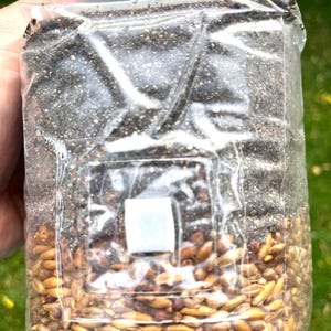 May include: A clear plastic bag filled with a dark, granular substance and grains. The bag is held by a hand, with a green grassy background visible. The bag's contents are divided, with grains at the bottom and the dark substance above.