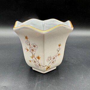May include: Cream-colored ceramic vase with a scalloped rim and a gold trim. The vase features a floral design with white blossoms and brown stems. The vase has a square base and a flared body.