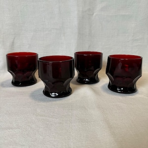 May include: Four ruby red drinking glasses. Each glass has a thick, faceted base and a cylindrical bowl. The glasses are made of a deep red glass, and the light reflects off the surfaces. The glasses are arranged on a neutral background.