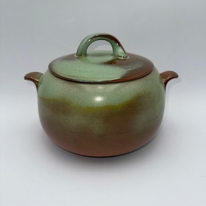 May include: A ceramic lidded pot with a rounded shape. The pot is glazed in shades of green, brown, and yellow. It has a handle on the lid and two side handles. The pot is likely used for cooking or serving food.