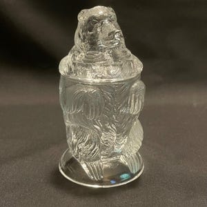 May include: A clear glass bear-shaped jar with a lid. The jar features a detailed bear design, including the head, body, and paws. The jar sits on a round base. The glass is transparent, allowing light to pass through.