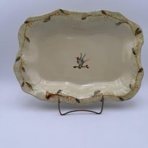 May include: Cream-colored rectangular ceramic serving dish with a scalloped edge. The rim is decorated with a leaf and berry design. A small bird illustration is centered on the dish. The dish is displayed on a metal stand.
