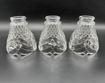 May include: Three clear glass lamp shades with a diamond pattern. The shades have a flared shape and a smooth rim. The glass is transparent, allowing light to pass through. The shades are likely for a chandelier or other type of light fixture.