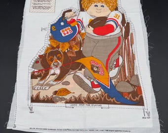 May include: A white fabric panel with a Cabbage Patch Kids design. The illustration shows a child in a football uniform with a football and a dog. The design includes a helmet, a pennant, and the text "Leave Open for Stuffing".