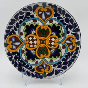May include: A decorative ceramic plate with a vibrant, hand-painted design. The plate features a central pattern of orange, blue, green, and brown floral motifs, surrounded by a border of blue leaf-like shapes. The plate is displayed on a clear stand.