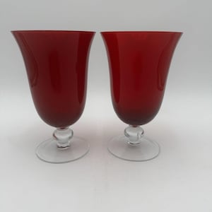 May include: Two matching stemmed glasses featuring deep red, flared bowls and clear glass bases. Each glass has a smooth, rounded stem connecting the vibrant red upper section to a circular, transparent foot, creating a balanced silhouette.