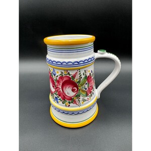 May include: A white ceramic mug with a large handle, decorated with red floral designs, green leaves, and blue and yellow accents. The mug has a yellow rim and base, and a green accent on the handle.