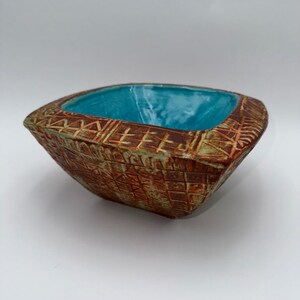 May include: A square ceramic bowl with a textured, geometric pattern in shades of brown and orange. The interior is a vibrant turquoise color. The bowl has a slightly rounded shape and is likely handmade.