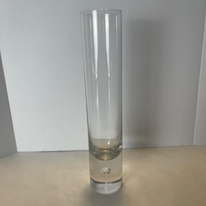 May include: A tall, clear glass vase with a cylindrical shape. The vase has a slightly rounded base with a small bubble detail. The glass is transparent, allowing light to pass through, and is suitable for displaying flowers or decorative stems.