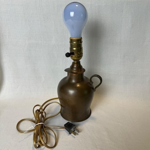 May include: A vintage bronze-colored lamp with a blue light bulb. The lamp has a round base with a handle and a brass-colored light fixture. The electrical cord and plug are visible. The lamp is on a white surface.