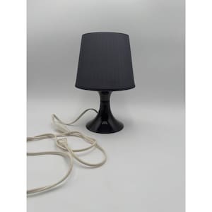 May include: A black table lamp with a lampshade and a base. The lampshade is a dark gray color, and the base is black. The lamp has a white power cord. The lamp is on a white surface.
