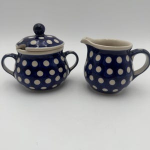 May include: A ceramic sugar bowl with a lid and a creamer. Both pieces are navy blue with white polka dots. The sugar bowl has two handles and a small spoon. The creamer has a handle and a spout.