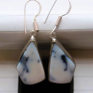 May include: A pair of drop earrings featuring irregular, teardrop-shaped white stones with black dendritic inclusions. The stones are set in silver-toned metal bezels and attached to silver-toned wire hooks with a coiled decorative detail.