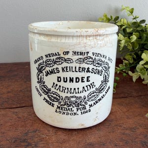 May include: A vintage white ceramic marmalade jar with a black label. The label reads "JAMES KEILLER & SONS DUNDEE MARMALADE" and includes text about awards and location. The jar has a slightly aged, crackled appearance.