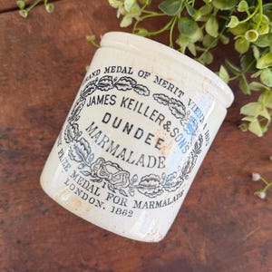 May include: An antique, off-white ceramic marmalade jar. The jar features a black and white label with the text "JAMES KEILLER & SONS DUNDEE MARMALADE" and "PRIZE MEDAL FOR MARMALADE LONDON. 1862". The jar is slightly aged, with a rustic appearance.