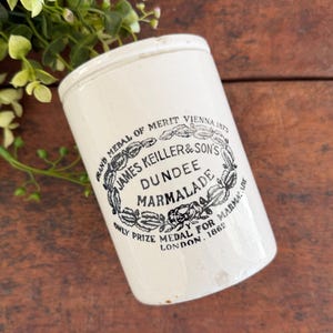 May include: Cream-colored antique marmalade jar with a lid. The jar features black text that reads "JAMES KEILLER & SON'S DUNDEE MARMALADE" and "ONLY PRIZE MEDAL FOR MARMALADE LONDON, 1862". The jar is cylindrical and has a slightly aged appearance.