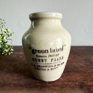 May include: A vintage, off-white ceramic jar with a rounded body and a narrow neck. The jar features the text "green label" and "Genuine INDIAN CURRY PASTE" in dark brown lettering. The jar is from L.A. Bearwood & Co. Ltd, London & Pont.
