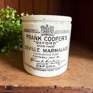 May include: A white ceramic jar with black printed text reading Frank Cooper's Oxford Seville Marmalade, featuring a royal crest, the phrase By Royal Appointment to His Majesty King George V, and the signature of Frank Cooper Limited.