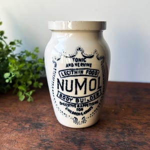 May include: Cream-colored ceramic jar with a slightly flared rim. The jar features a shield-shaped label with the text "TONIC AND NERVE, LECITHIN FOOD, NUMOL, BODY BUILDER, DIGESTIVE & APPETISING FOR CHILDREN & ADULTS" in dark blue lettering.