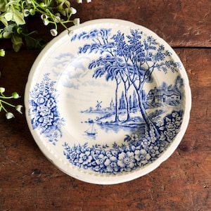 May include: A round ceramic plate featuring a blue and white transferware pattern. The design depicts a landscape with a sailboat, a windmill, trees, and a house, framed by floral borders. The plate has a scalloped edge and a cream-colored base.
