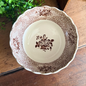 May include: A round ceramic bowl with a scalloped edge, featuring a cream-colored base and intricate brown transferware patterns. The design includes a central floral basket motif and a decorative border of scrolling vines and small flower baskets.