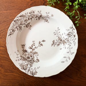 May include: A white ceramic plate with a scalloped edge, adorned with a brown floral pattern. The design features detailed botanical illustrations of flowers and leaves, creating an elegant, vintage aesthetic. Ideal for serving food.