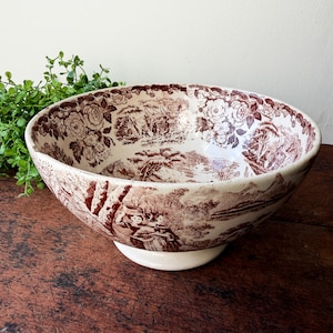May include: A large, antique serving bowl with a white base and a wide rim. The bowl features a brown transferware design of a pastoral scene with floral accents. The bowl is set on a dark wooden surface.