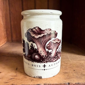 May include: A cylindrical off-white ceramic crock featuring a dark brown transferware illustration of two mushrooms and an acorn. The text "SOUS-BOIS" and "AUTOMNE" is printed in dark brown lettering near the base, separated by a fleur-de-lis symbol.