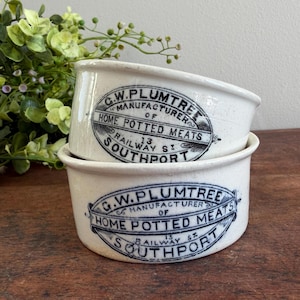 May include: Two antique white ceramic pots stacked on top of each other. Each pot has a black and blue oval label with the text "G.W. Plumtree Manufacturer of Home Potted Meats 13 Railway St Southport".