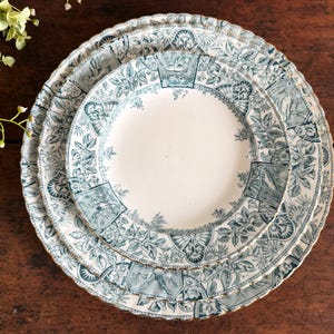 May include: A stack of three antique white plates with a teal floral and architectural design. The plates have scalloped edges and a gold trim. The plates are arranged on a dark wood surface.