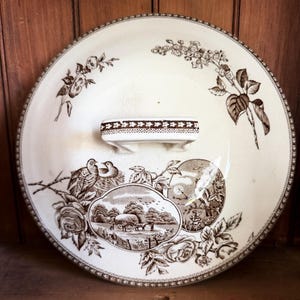May include: A round ceramic lid featuring a brown transferware pattern on a cream background. The design includes floral motifs, a central landscape scene with a horse-drawn cart, and a bird in a nest. It has a rectangular handle with a geometric border.