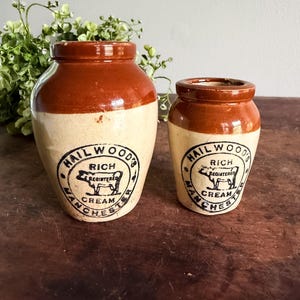 May include: Two antique cream jars with brown tops and off-white bodies. Each jar features a black circular label with the text "HAIL WOOD'S RICH REGISTERED CREAM MANCHESTER". The jars are displayed on a wooden surface.