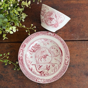 May include: A white ceramic teacup and saucer set with a red toile pattern. The teacup is angled, revealing the interior design. The saucer and teacup feature detailed illustrations of animals and landscapes, with a decorative border.