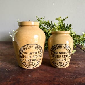 May include: Two antique cream jars with a tan glaze. Each jar has a black and white label that reads "Manchester Creamery Hailwood's Pure Rich Cream Broughton". The jars are different sizes and sit on a wooden surface.