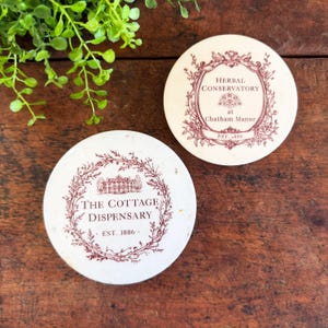 May include: Two round, off-white coasters with maroon text and illustrations. One coaster reads "The Cottage Dispensary, EST. 1886" and the other reads "Herbal Conservatory at Chatham Manor, EST. 1886". Both have a floral wreath design.
