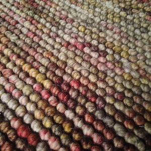 Può includere: Primo piano di un tappeto strutturato con un motivo di piccoli elementi arrotondati e colorati. Il tappeto presenta una sfumatura di colori, tra cui tonalità di rosso, rosa, marrone e beige, creando una superficie visivamente accattivante e tattile.