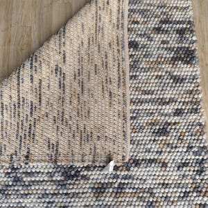 Puede incluir: Una alfombra texturizada con una paleta de colores neutros. La alfombra presenta una mezcla de tonos beige, gris y blanco, con un patrón en relieve punteado en la superficie superior. Se ve la parte inferior de la alfombra, mostrando una construcción tejida.
